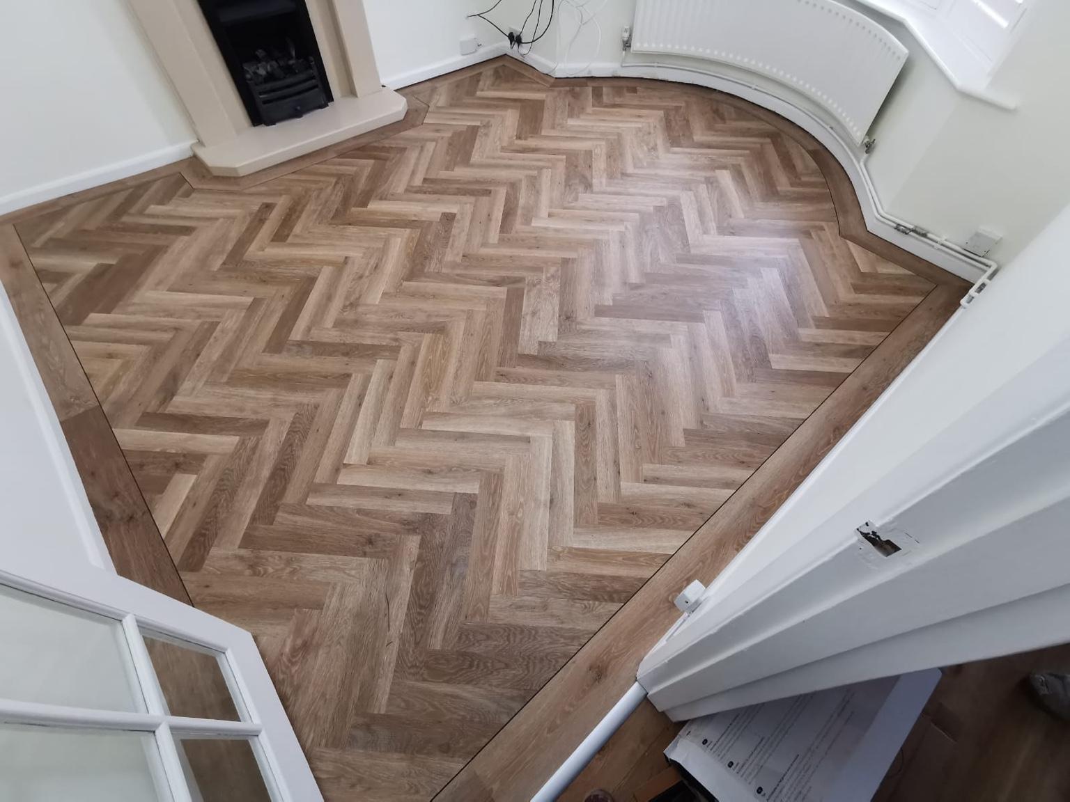 A completed living room floor: herringbone flooring with a perimeter border cut to follow the curve of the bay window, running past the fireplace